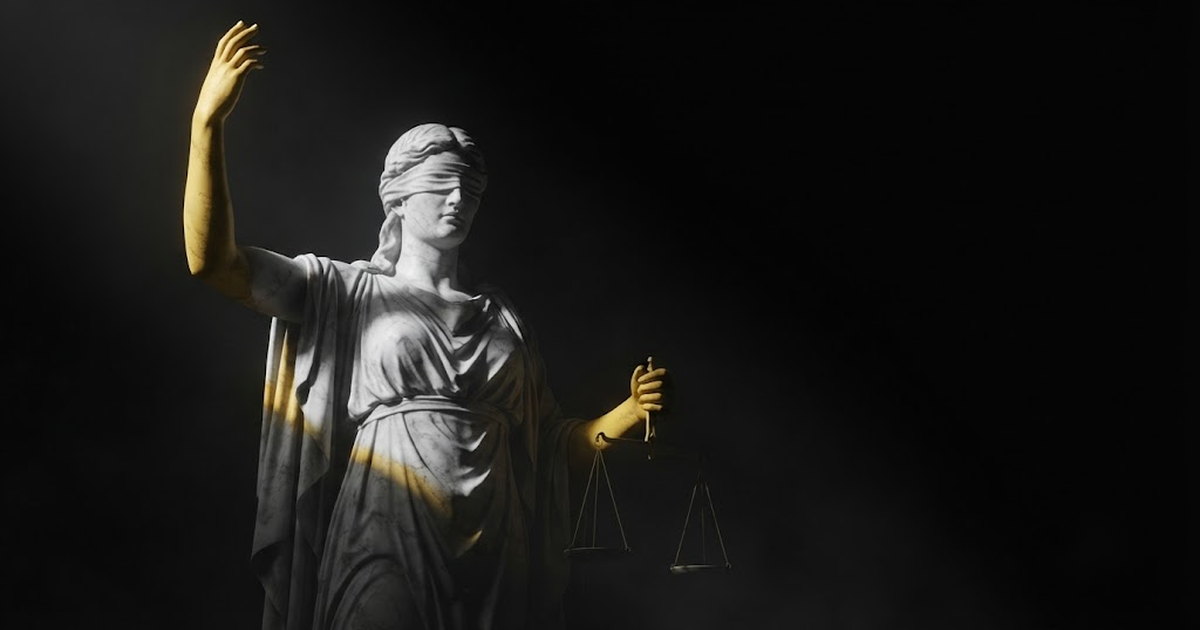 Colossal blindfolded marble statue of Themis with one hand raised in judgement, lit by a single blade of gold light